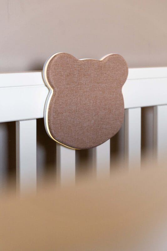A wooden crib displays a Leilas Bear White Noise Night Light, featuring a brown fabric-covered bear head plaque on the headboard. The blurred background emphasizes this decorative element.