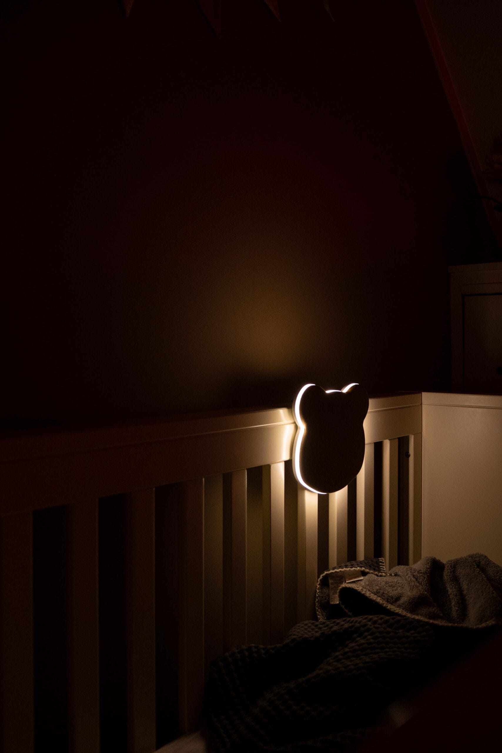 A dimly lit room with Leilas Bear White Noise Night Light attached to a cribs railing. Its soft glow casts warmth over a dark blanket draped inside, creating a cozy atmosphere.