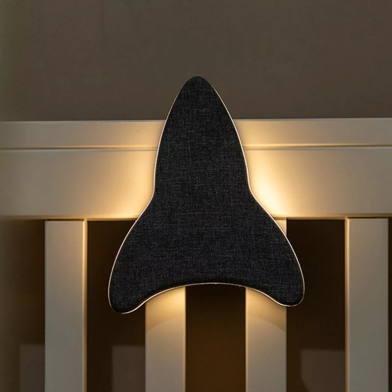 Lifestyle foto van Ollie & Leila's Ollie's Rocketship, een grijs raketvormig nachtlampje, bevestigd aan een babybedje in een schemerige kamer, dat een zachte, warme gloed uitstraalt voor een kalmerende nachtrust.
