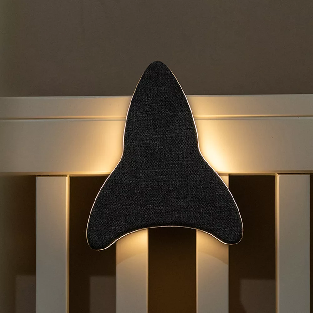 Lifestyle foto van Ollie & Leila's Ollie's Rocketship, een grijs raketvormig nachtlampje, bevestigd aan een babybedje in een schemerige kamer, dat een zachte, warme gloed uitstraalt voor een kalmerende nachtrust.