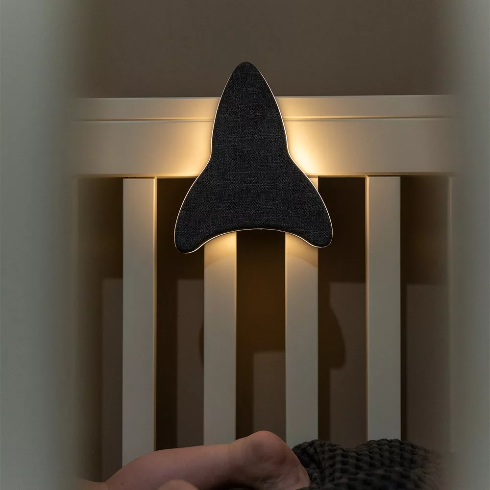 Sfeerfoto van Ollie & Leila's Ollie's Rocketship nachtlampje, dat een zachte gloed verspreidt aan het bedje van een slapend kind in een donkere kamer, en zo een veilige en rustgevende slaapomgeving creëert.