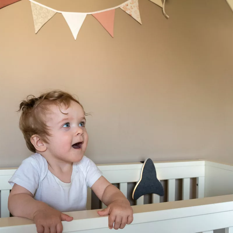 Blije baby in een wit wiegje kijkt verwonderd naar Ollie & Leila's Ollie's Rocketship, een grijs raketvormig nachtlampje en witte ruis machine, voor een rustgevende sfeer in de babykamer.