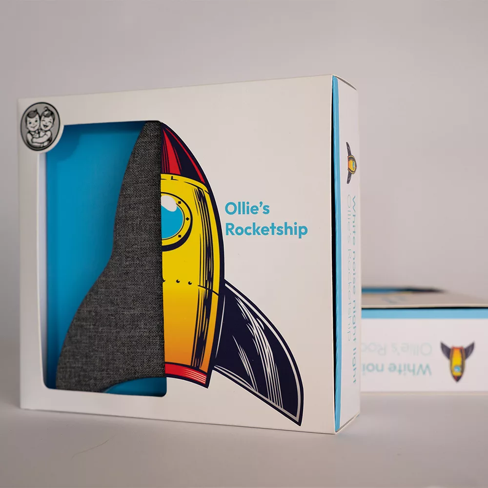 Geschenkverpakking van Ollie & Leila's Ollie's Rocketship, een wit met blauw doosje met een kijkvenster dat het raket nachtlampje en een kleurrijke illustratie van de raket toont, ideaal als babycadeau.