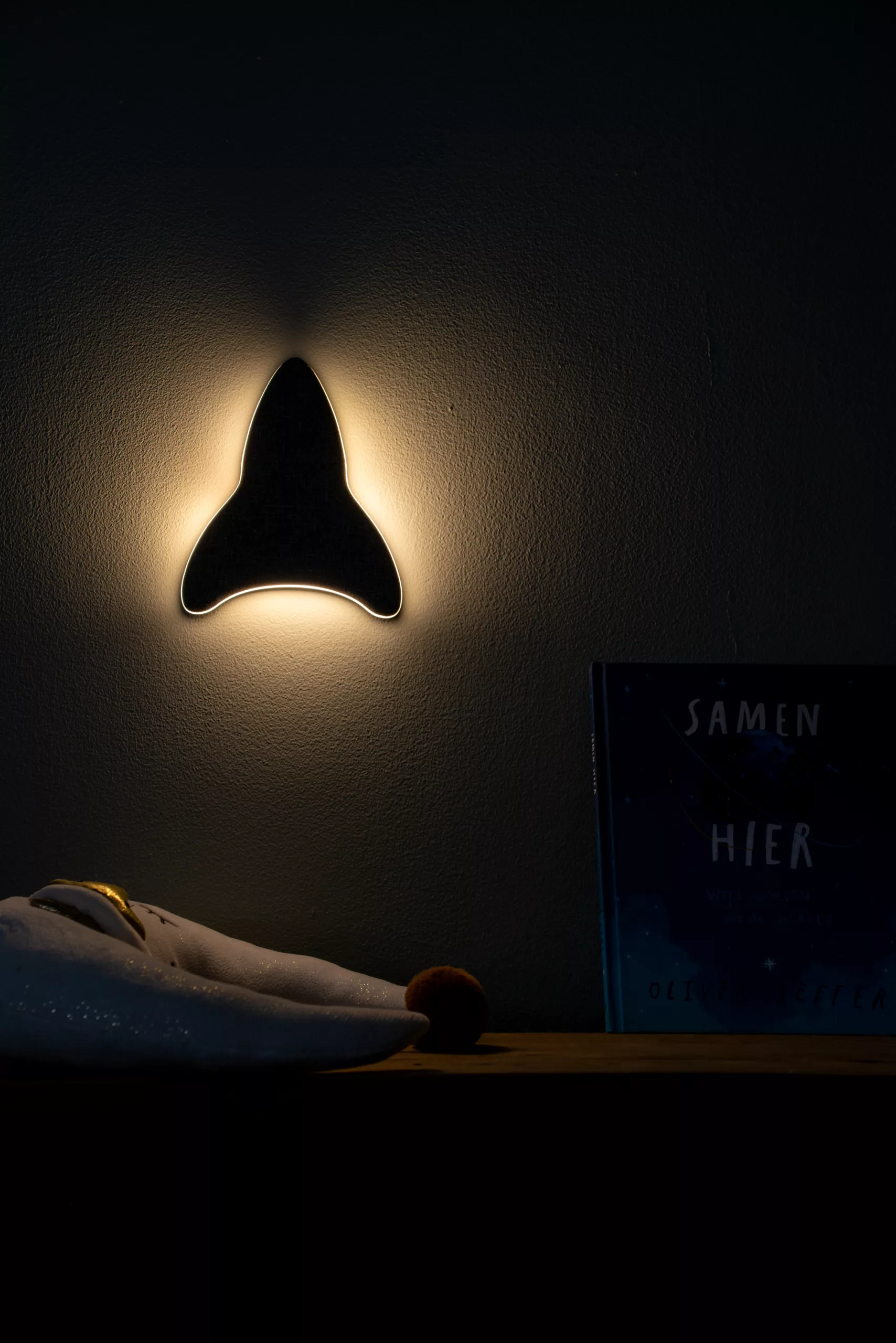 Sfeervolle opname van Ollie & Leila's Ollie's Rocketship, een raketvormig nachtlampje, gemonteerd aan de muur in een kinderkamer, dat een warme, uitnodigende gloed verspreidt boven een plank met een knuffel en het boek "SAMEN HIER".